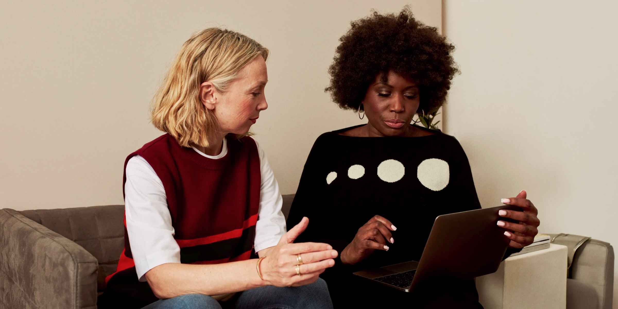 Two women sit together on a grey sofa in a relaxed office setting, one gesturing as she talks while the other reviews something on a laptop.