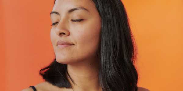 Close-up of a person with medium-length dark hair, eyes closed and a peaceful expression, set against a bright orange background