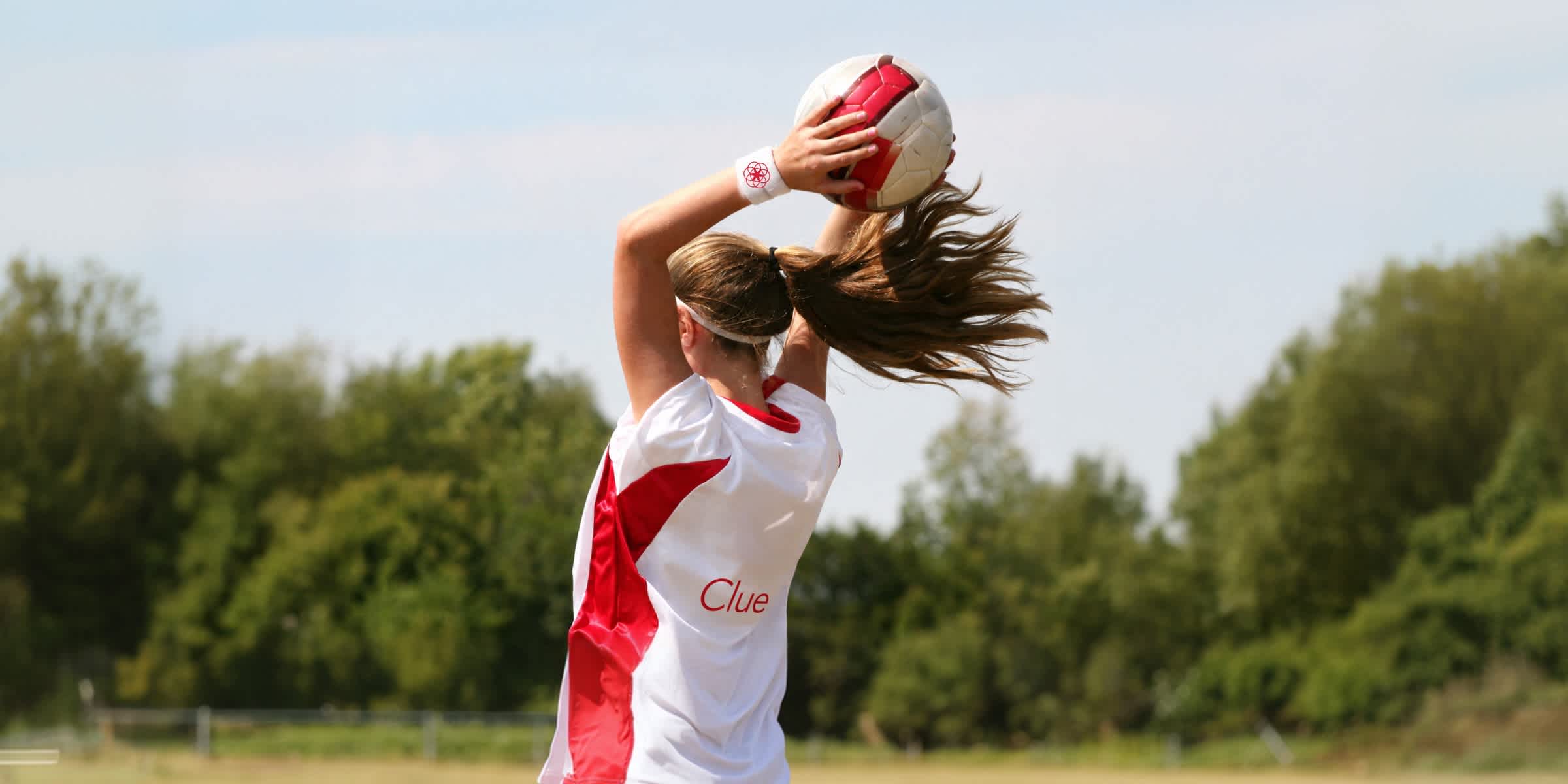 woman holding a football in both hands above her head and wearing a white t-shirt with the Clue logo on the back