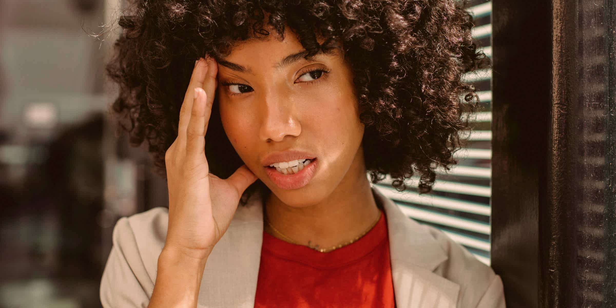 A woman with curly hair presses a hand to her temple, glancing to the side with a strained expression.
