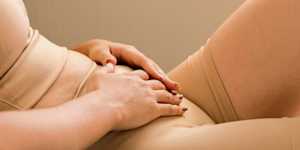 Person in beige underwear and a crop top resting both hands on their lower abdomen.