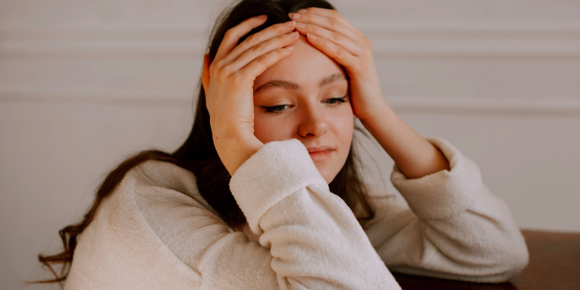 A young woman with long dark hair rests her elbows on a wooden table, both hands pressed to her forehead, looking down with a stressed, overwhelmed expression.