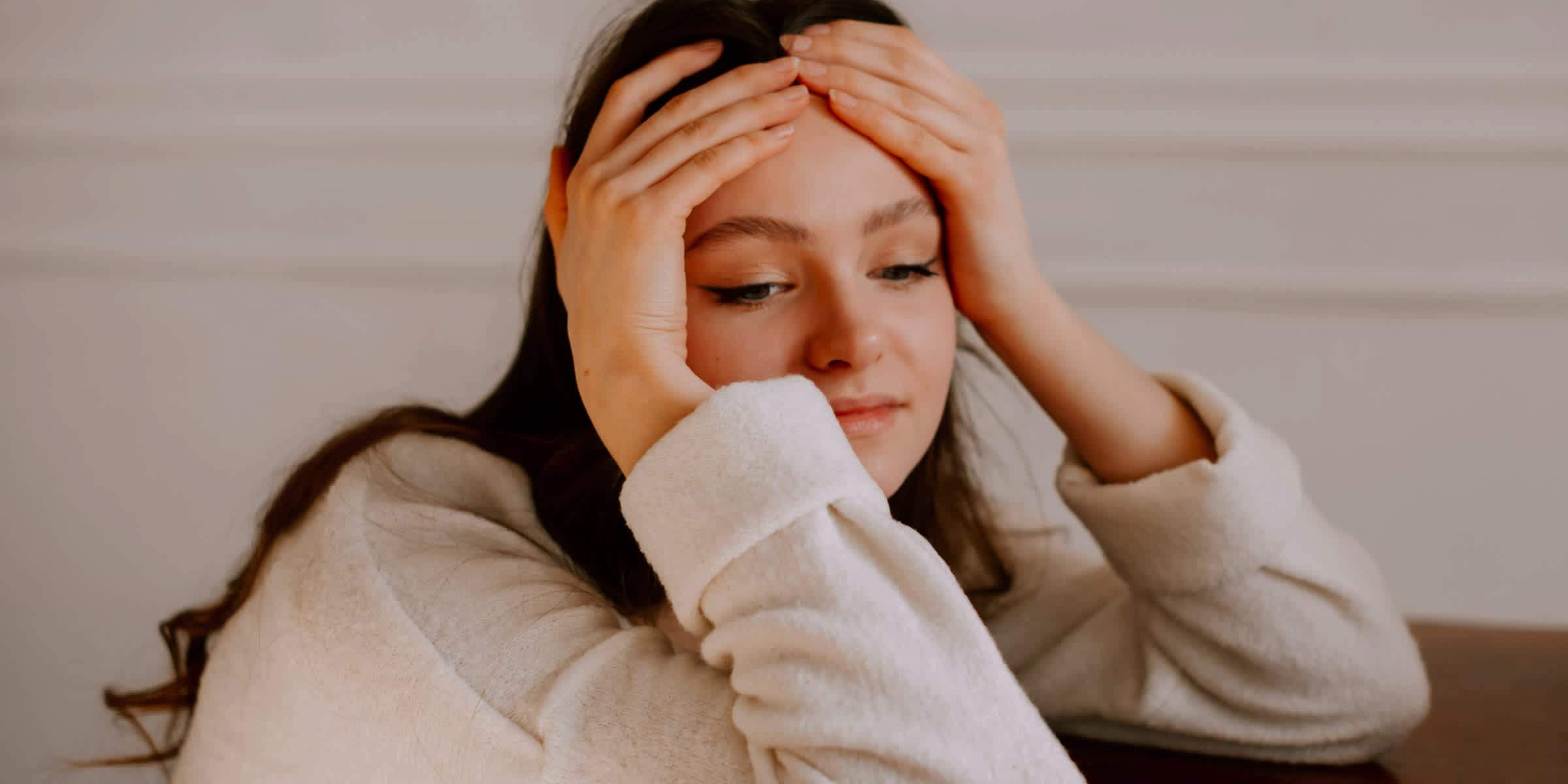 A young woman with long dark hair rests her elbows on a wooden table, both hands pressed to her forehead, looking down with a stressed, overwhelmed expression.