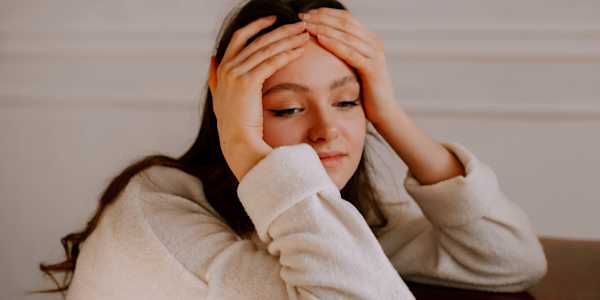 A young woman with long dark hair rests her elbows on a wooden table, both hands pressed to her forehead, looking down with a stressed, overwhelmed expression.