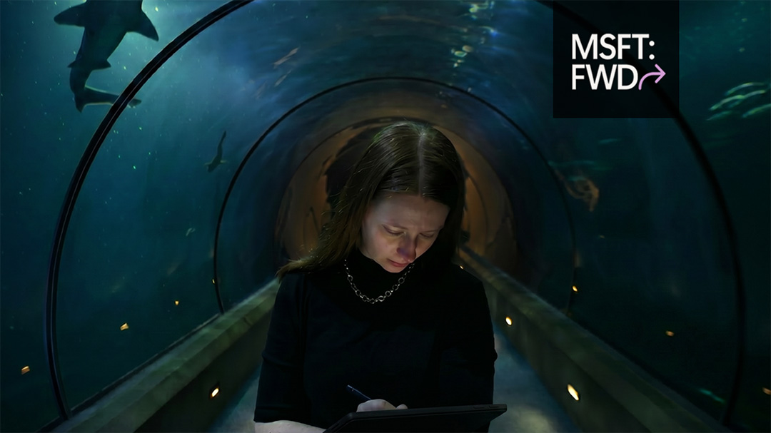 Person using a Surface device with a pen inside an underwater tunnel aquarium, with sharks swimming above and around.