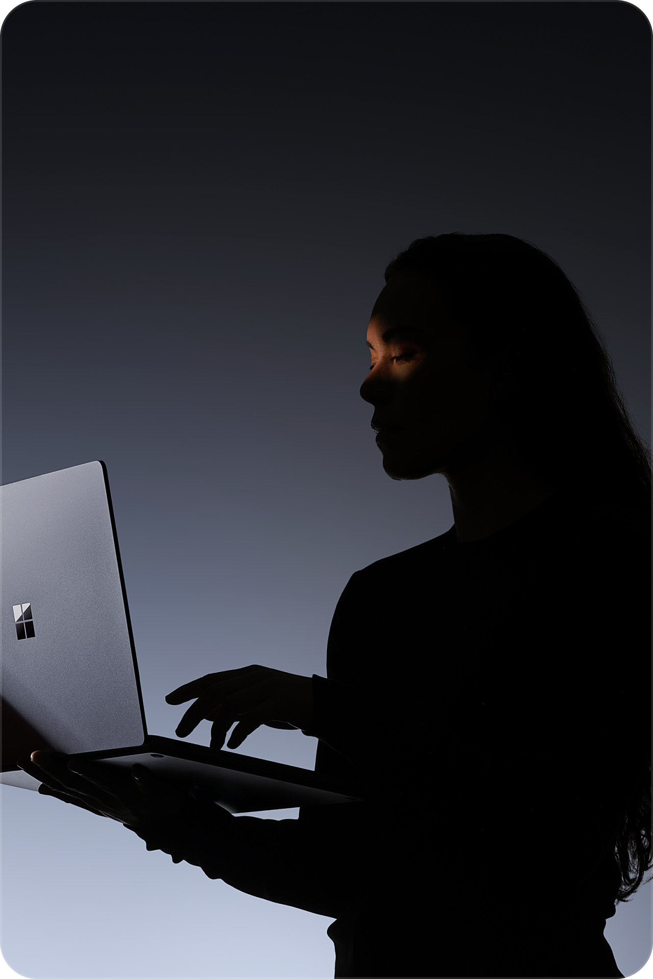 Silhouette of a person using a Microsoft Surface laptop against a dark background with soft backlighting