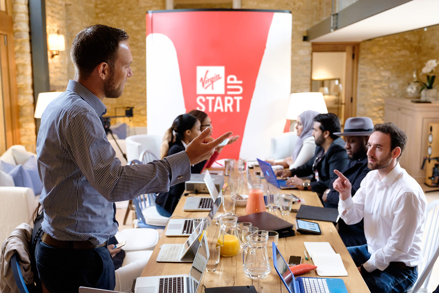 Seven people around a long table having a meeting, one person is standing up talking