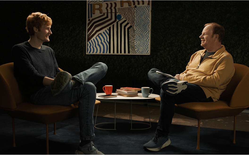 Image of Cursor CEO Michael Truell on the left and Rippling CEO Parker Conrad on the right. Both are seated across from eachother in a makeshift living room set and they are both talking candidly