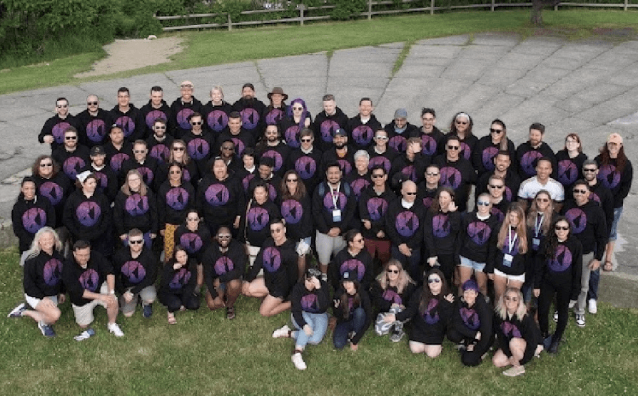 Large group gathered outdoors wearing matching black shirts with purple circular logos, posed together on grass.