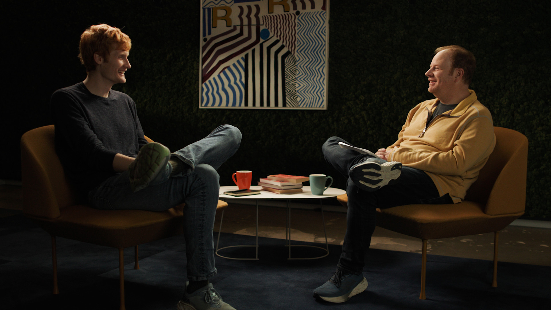 Photograph of Michael Truell, CEO of Cursor, and Parker Conrad, CEO of Rippling, smiling at each other while sitting in midcentury modern style chairs in a room with studio lighting