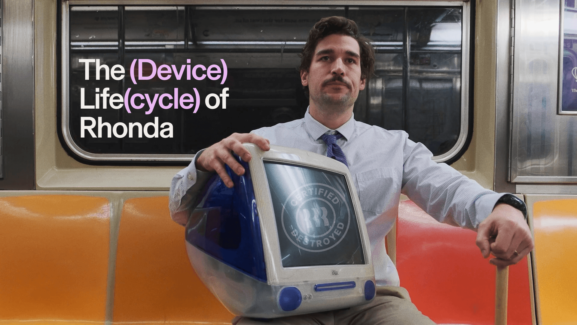 A person in office clothes sits on an orange subway seat holding a bulky blue-and-white vintage desktop computer in his lap. Large text on the subway window reads, “The (Device) Life(cycle) of Rhonda." The computer screen shows a circular stamp that says “Certified Destroyed.”