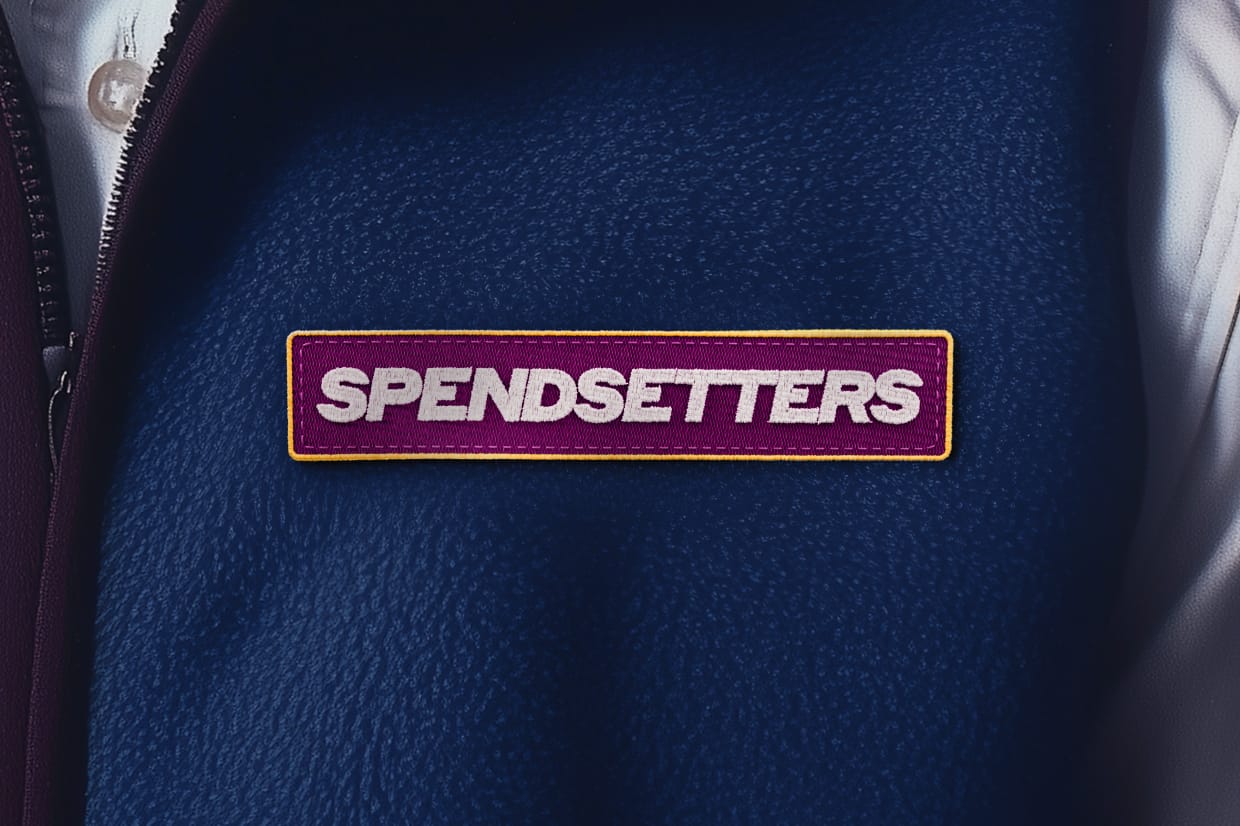Close-up of a dark blue textured garment with a burgundy and yellow embroidered patch. The patch displays the word "SPENDSETTERS" in bold white letters.