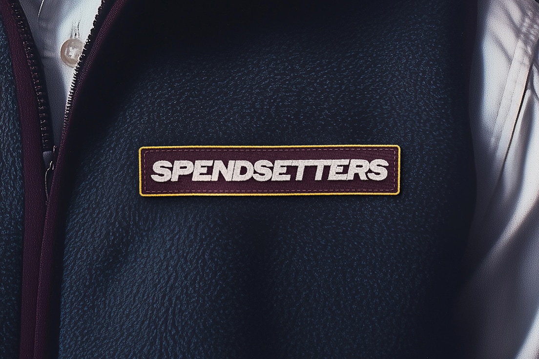 Close-up of a dark blue textured garment with a burgundy and yellow embroidered patch. The patch displays the word "SPENDSETTERS" in bold white letters.