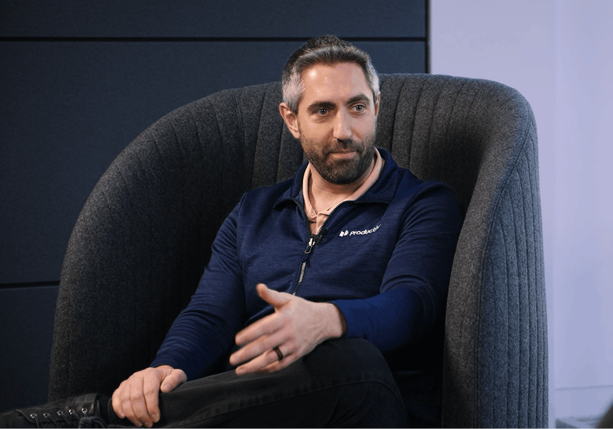 A person in a navy blue polo shirt seated in a gray high-backed chair against a dark wall during an interview.
