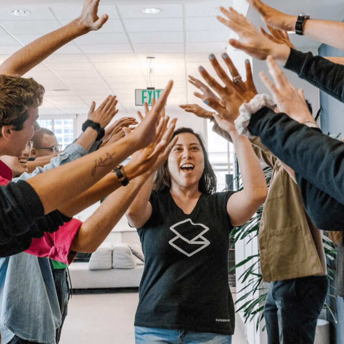 Enthusiastic group celebrating with raised hands in an office space, woman in black shirt with logo at center.