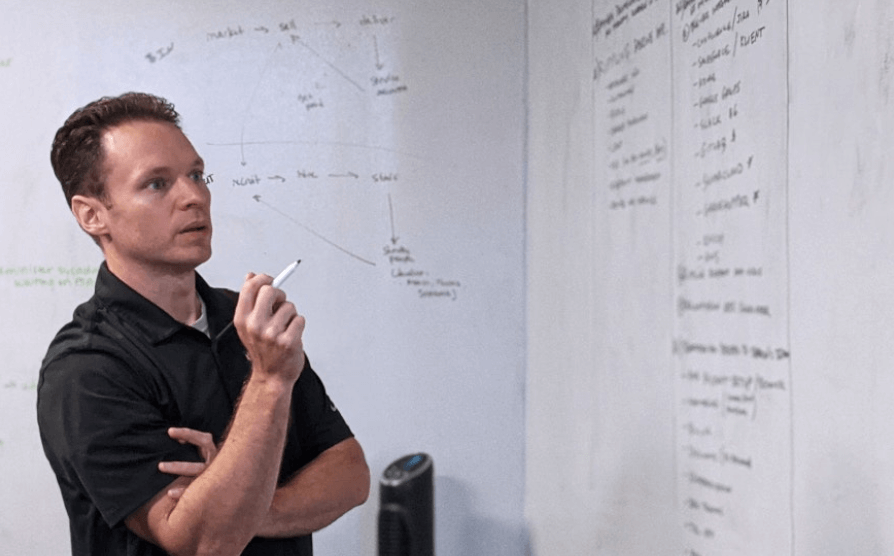 Person in black shirt holding marker pen while looking at whiteboard with flowcharts and business notes.