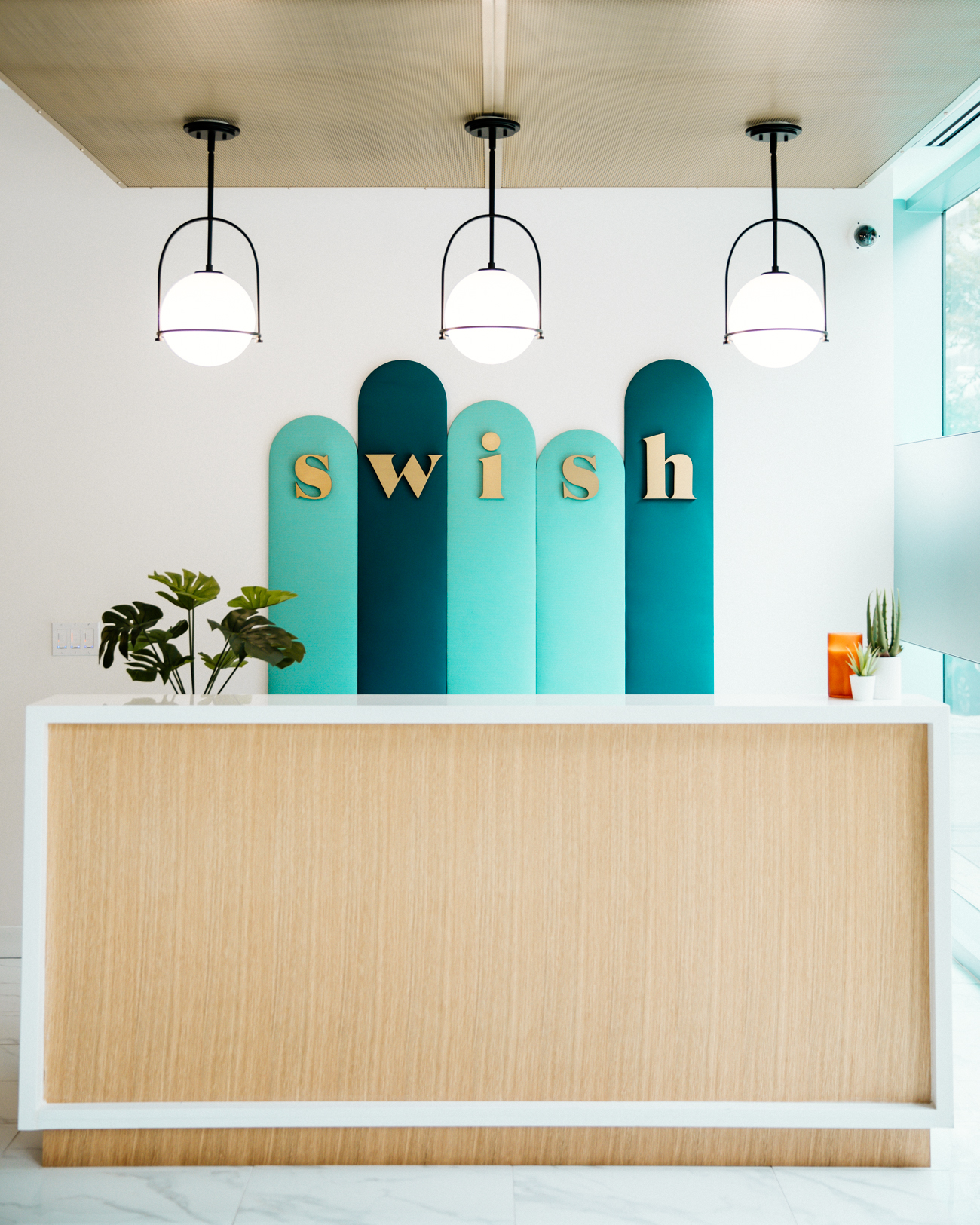 Modern reception desk with "swish" gold lettering on teal arched panels, pendant lights above, and potted plants.