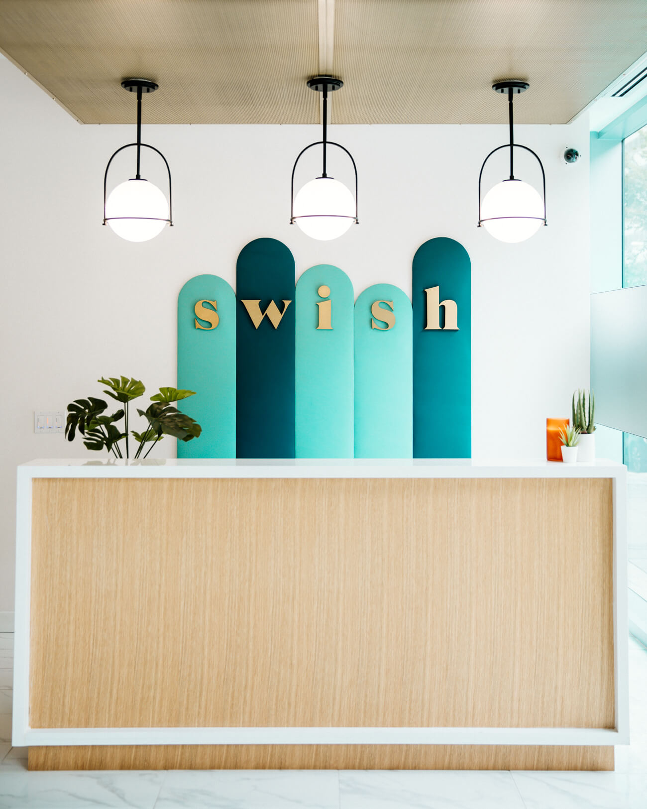 Modern reception desk with “swish” gold lettering on teal arched panels, pendant lights above and potted plants.