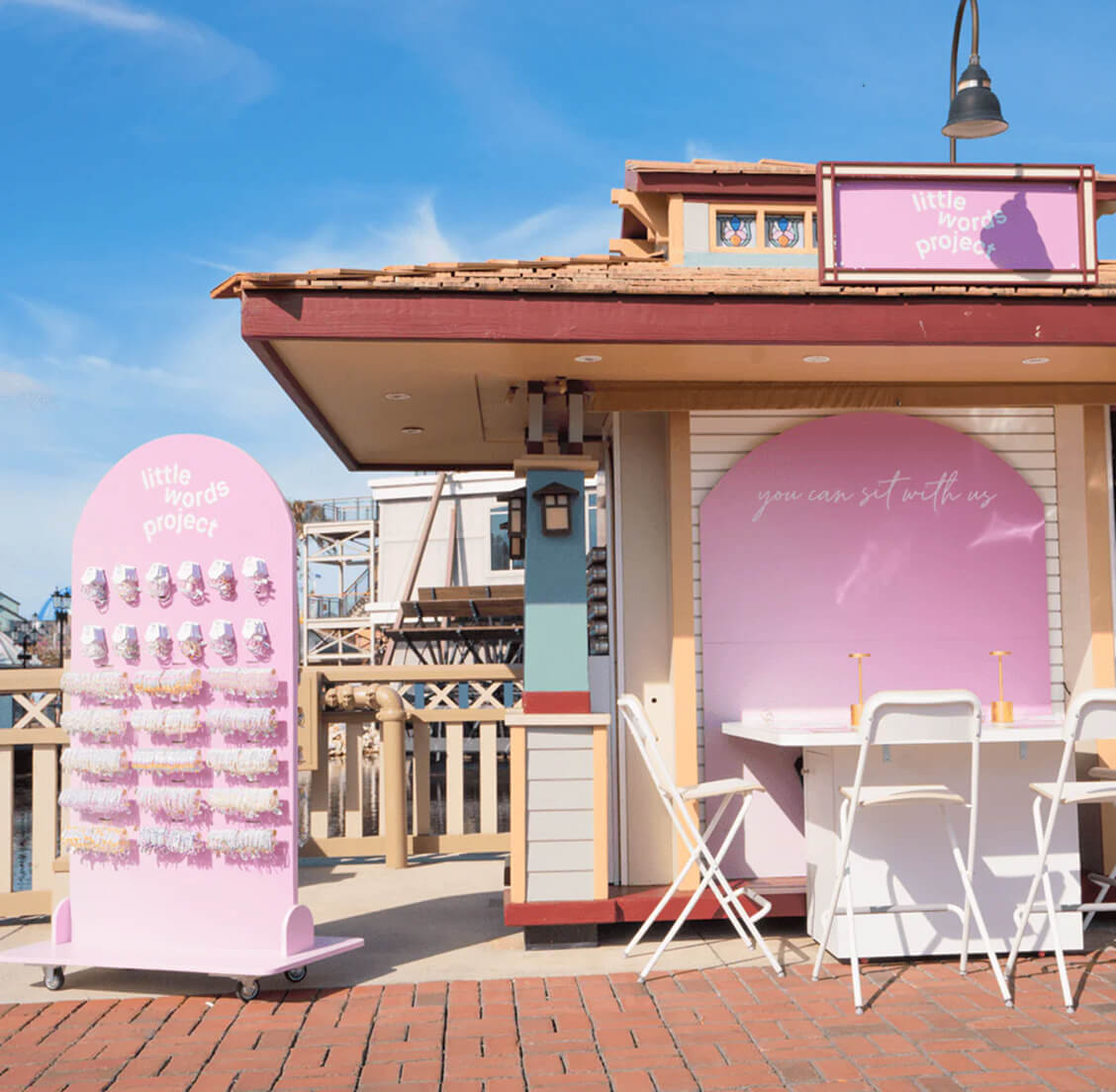 Pink-themed “Little Words Project” retail kiosk with bracelet display and outdoor seating area under blue sky.