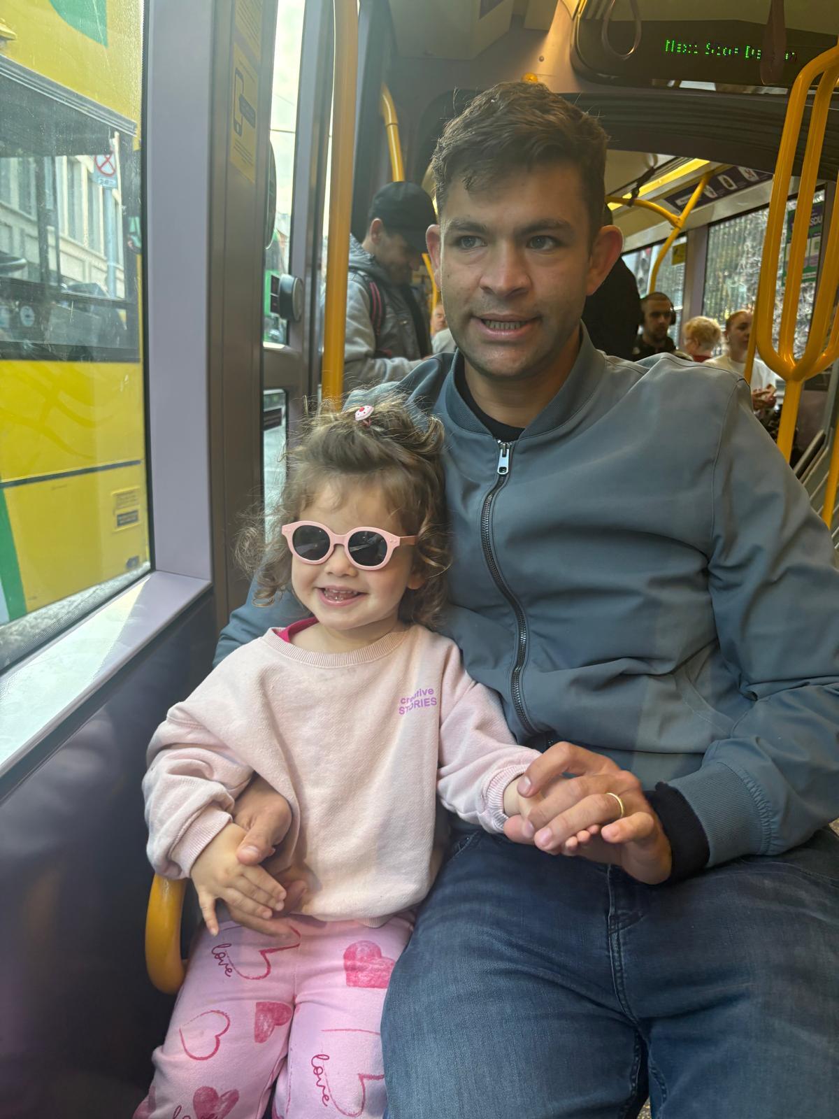 A young child wearing pink sunglasses and pink outfit sitting next to an adult on public transportation.