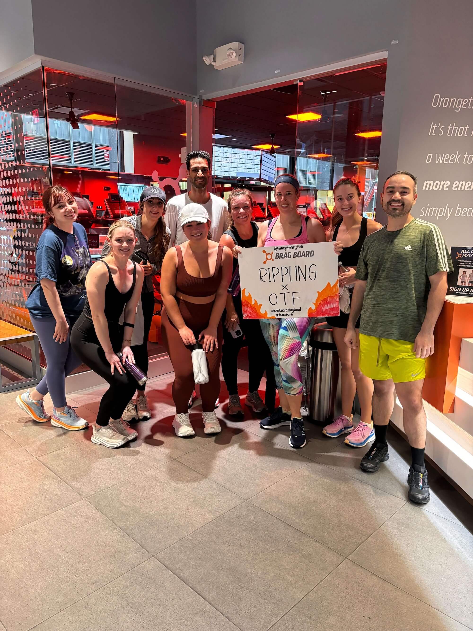 Geo and members of Orange Theory Fitness posing with a poster that says "Brag board: Rippling x OTF"
