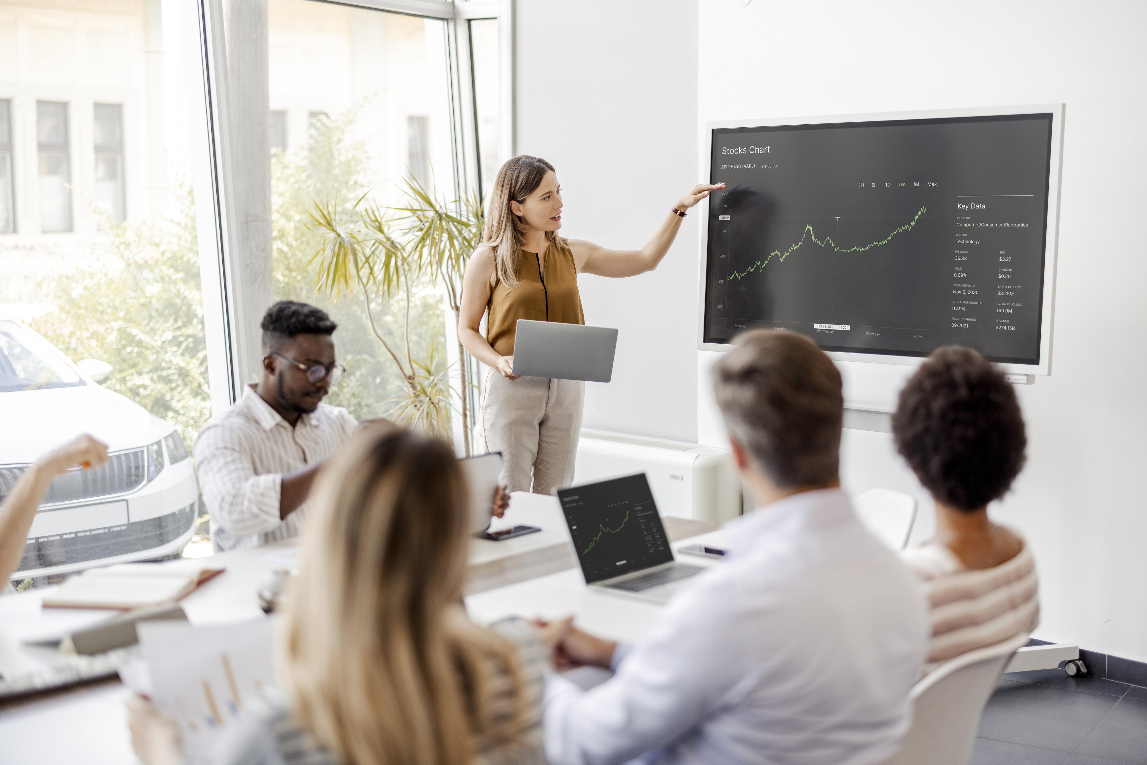 Demand Analysis & Forecasting - group of people in a meeting room with a female presenter gesticulating to a screen showing a forecast diagram.
