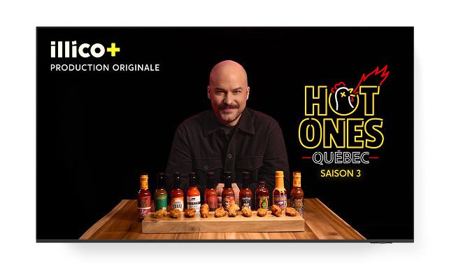 A man sits behind a row of hot sauce bottles and wings for "HOT ONES QUÉBEC SAISON 3" on illico+.
