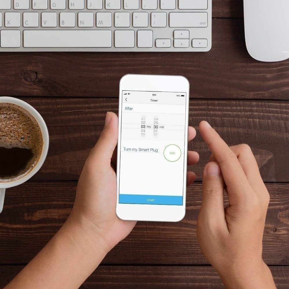 Hands using smartphone to control smart home devices, with coffee and keyboard on wooden desk.