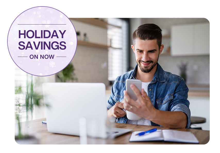 A man sitting in front of a laptop and viewing his smartphone. A roundel on the left reads “Holiday Savings on now”.