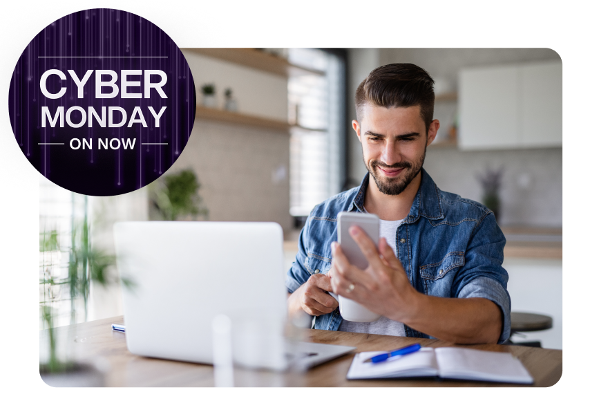 A man sitting in front of a laptop and viewing his smartphone. A roundel on the left reads “Cyber Monday on now”. 
