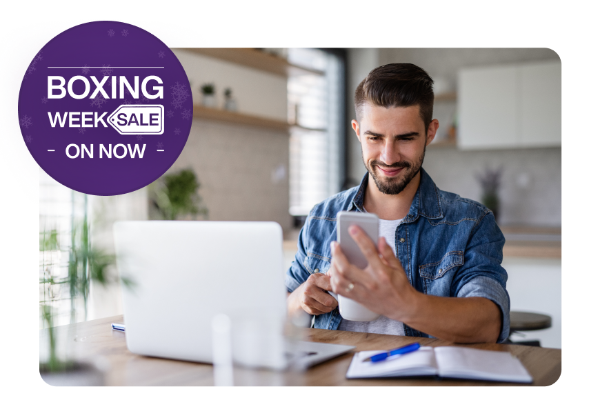 A man sitting in front of a laptop and viewing his smartphone. A roundel on the left reads “Boxing Week Sale on now”.