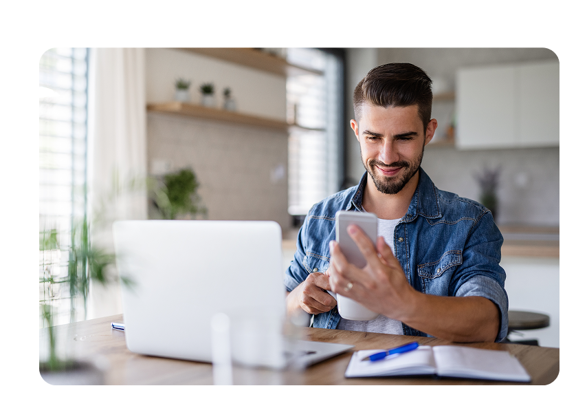 A man sitting in front of a laptop and viewing his smartphone.