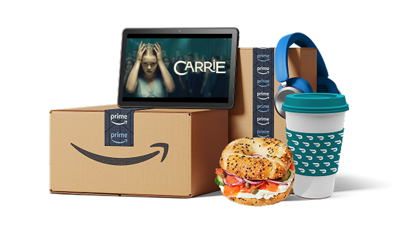 A tablet playing Carrie, Amazon Prime delivery boxes, blue headphones, a coffee cup, and a bagel sandwich.