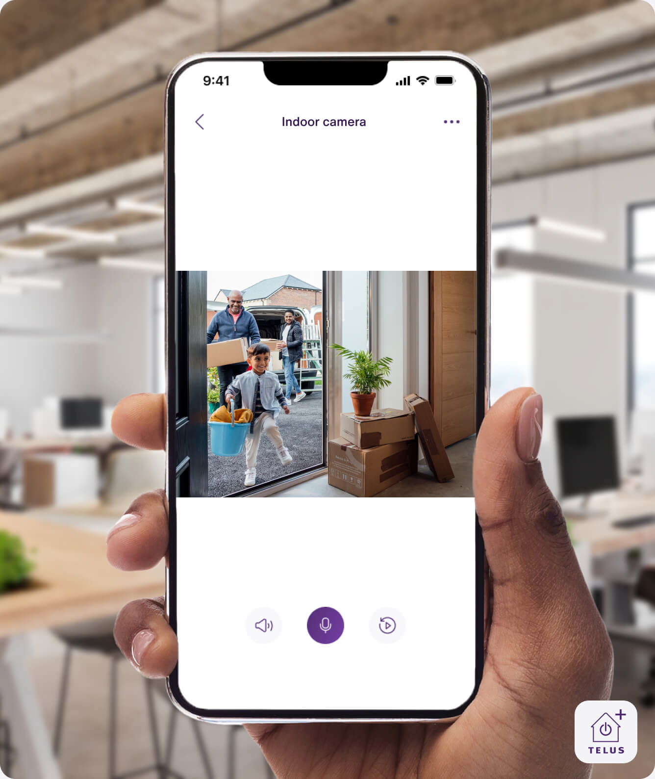 A hand holding the TELUS SmartHome+ app from a workplace setting shows family members coming into the house from the TELUS Indoor Camera.