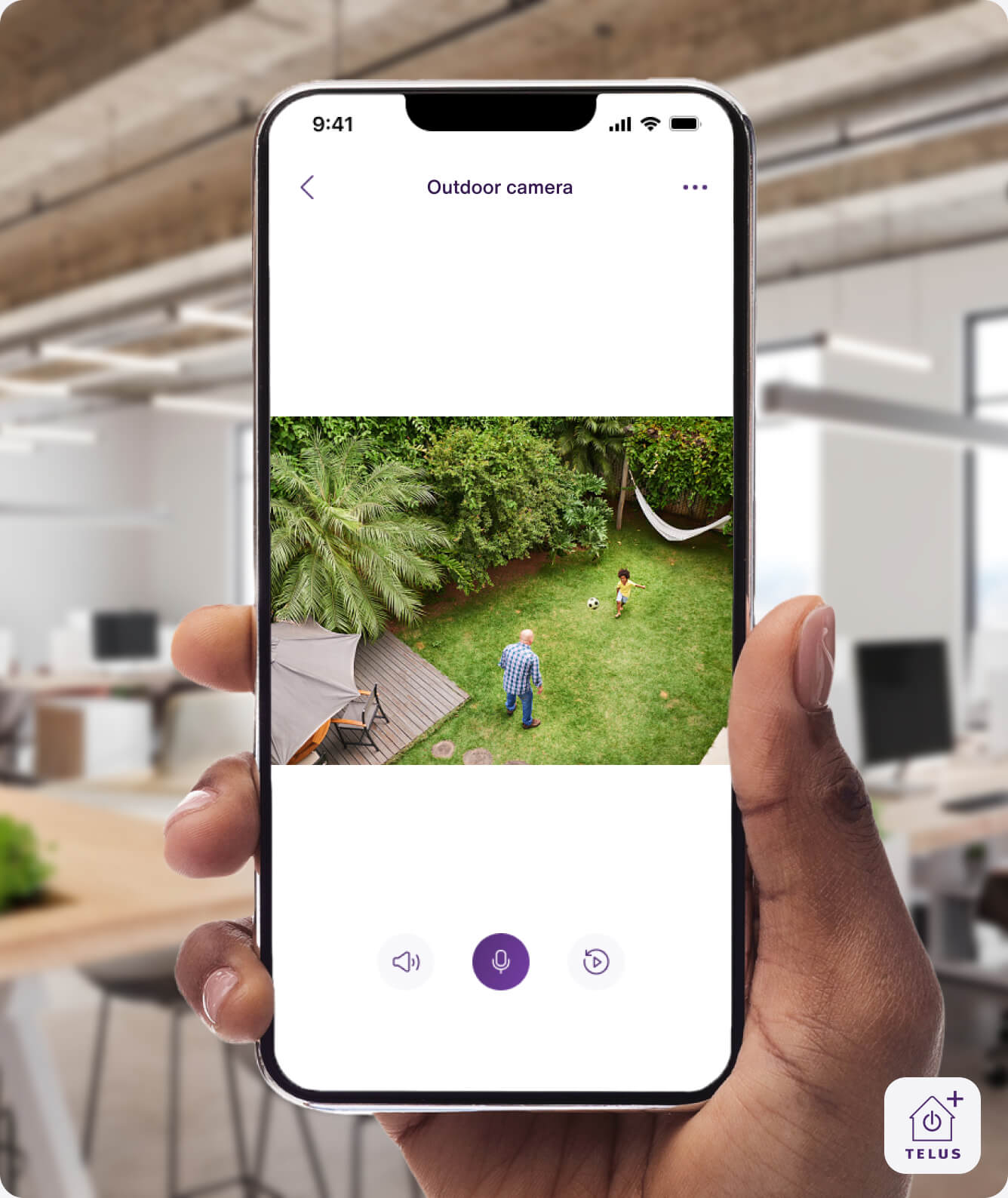 A hand holding the TELUS SmartHome+ app showing a family playing in the backyard from the TELUS Outdoor Camera.