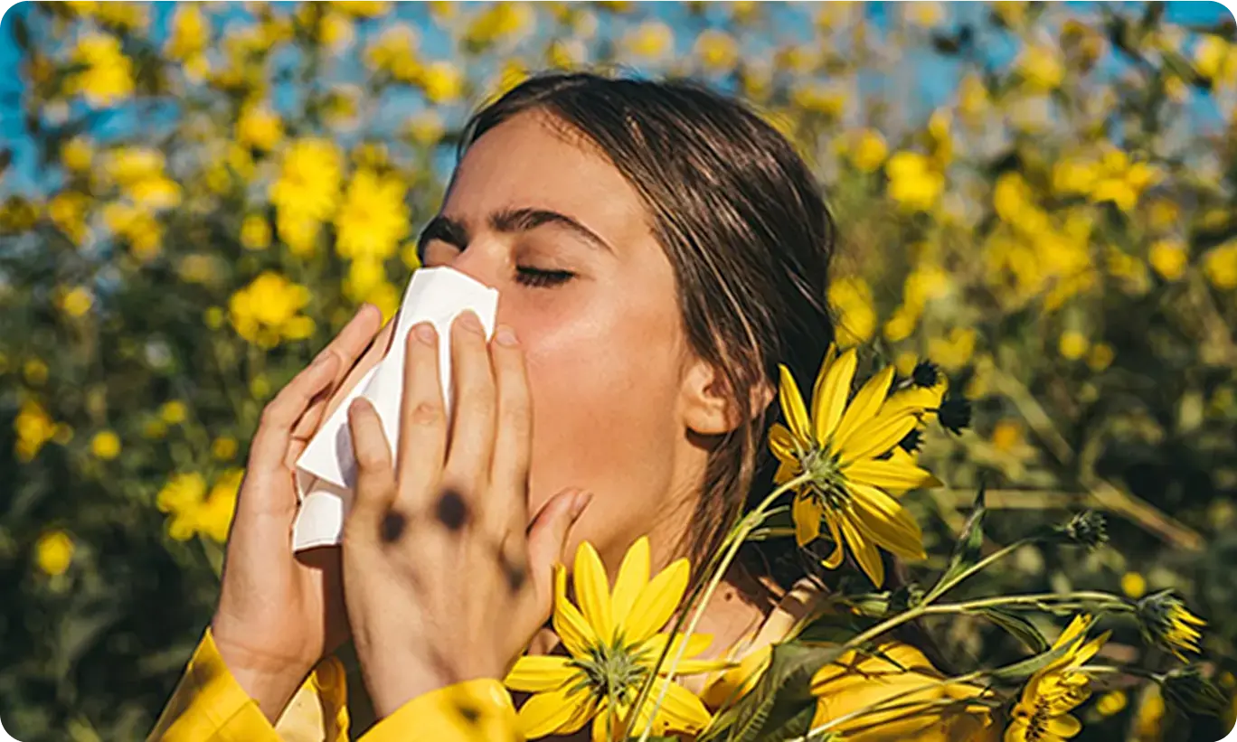 A woman holding a tissue in a field of yellow flowers, showing that allergies are in everyday life.