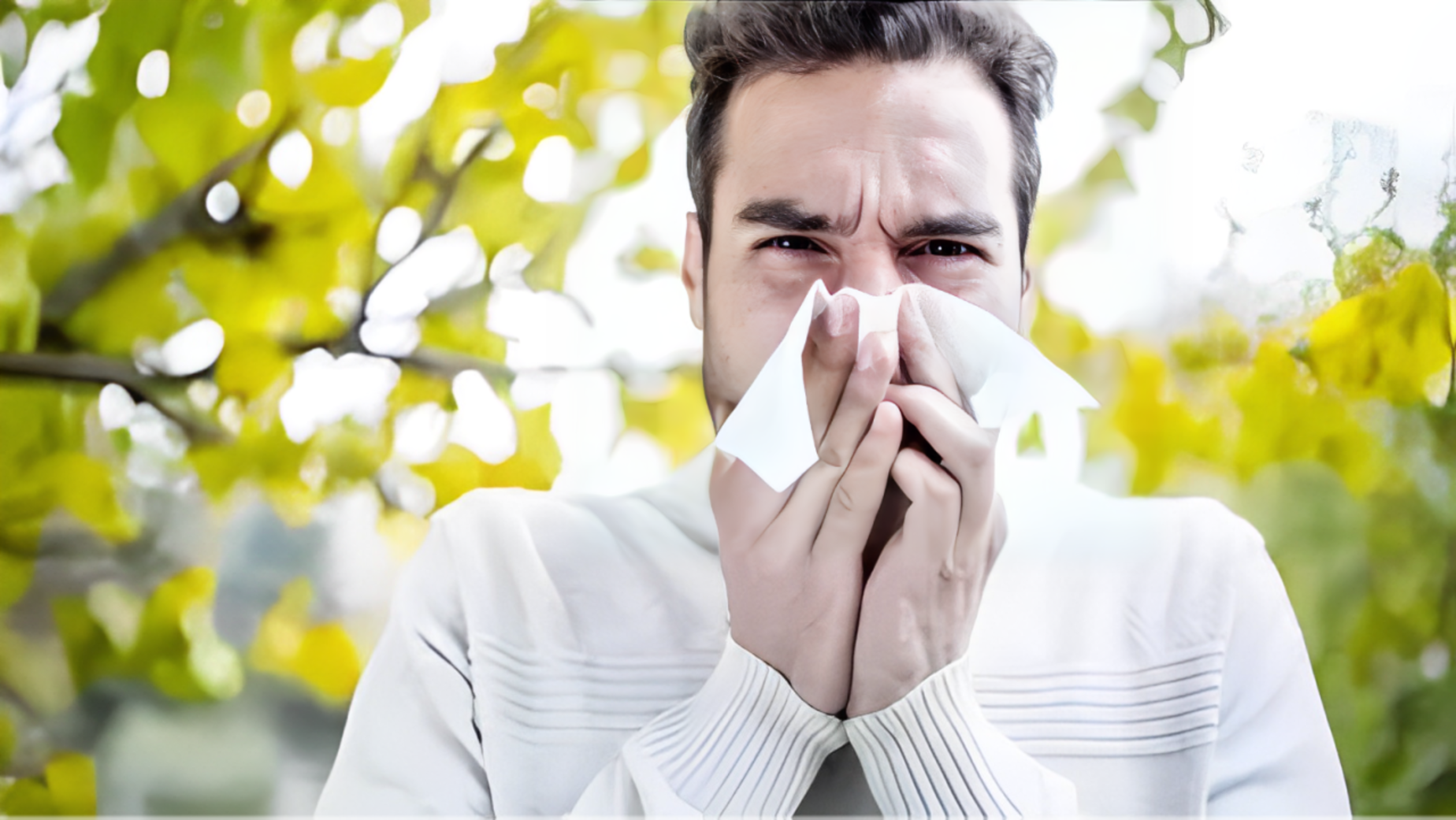 A man holding a tissue near his face, surrounded by foliage demonstrating the discomfort of seasonal allergies.