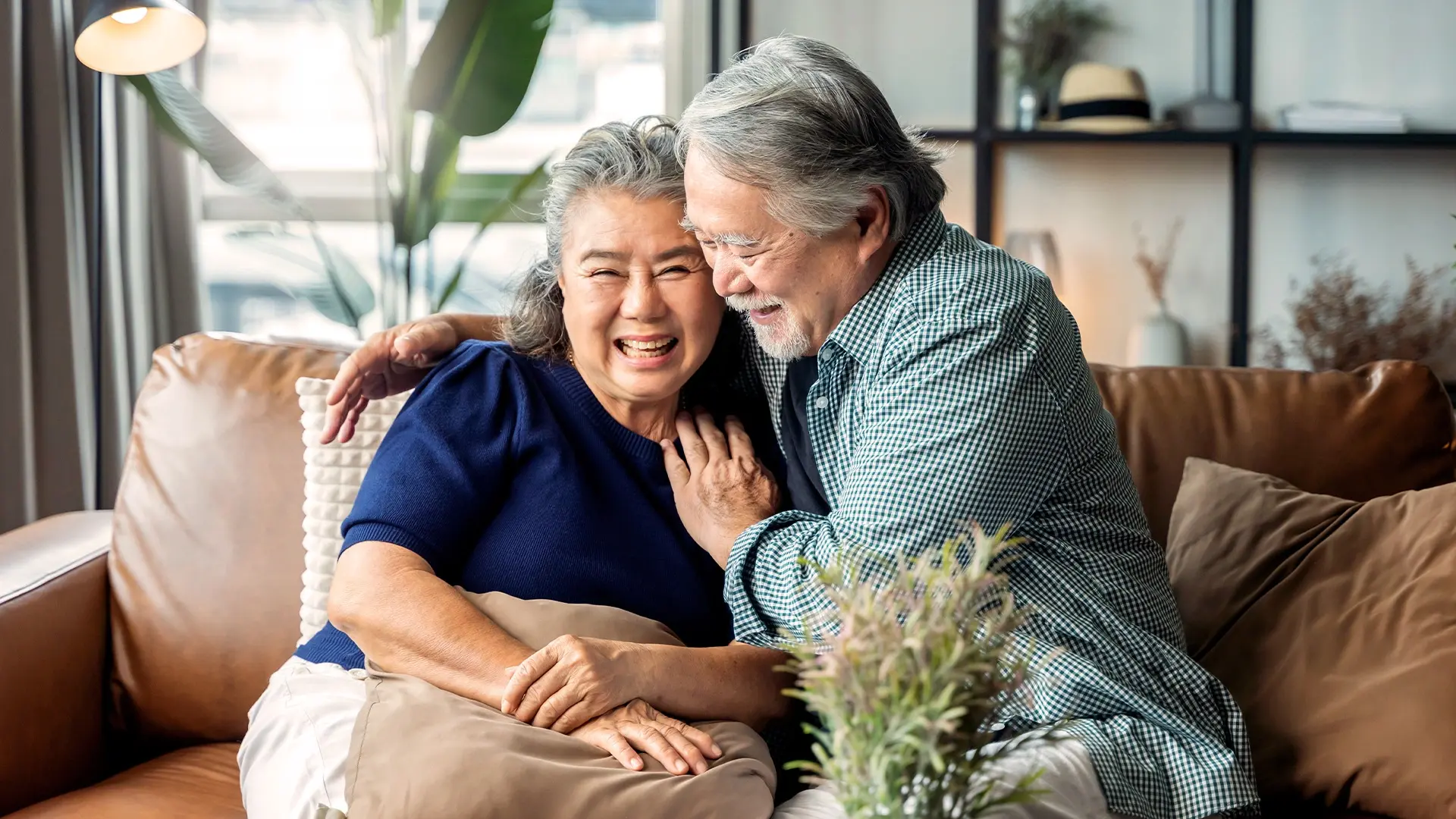  Elderly couple sitting on a brown leather sofa, smiling with the man in a blue checkered shirt and the woman in a navy shirt, surrounded by a lamp and houseplant.