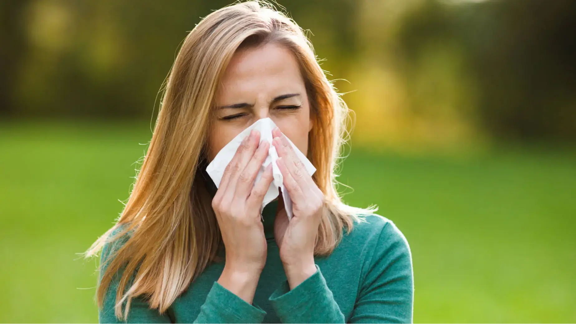 A woman holding a tissue near her face in a grassy outdoor setting, demonstrating the discomfort of allergy symptoms.de