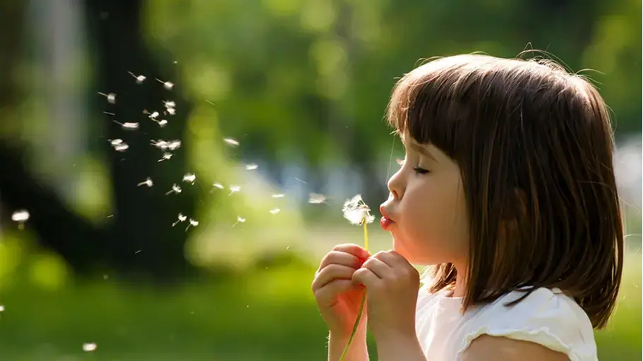 A young girl outside surrounded by trees blowing on a dandelion dispersing the seeds into the wind
