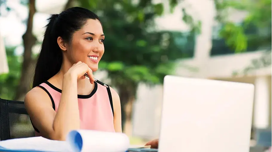 A young woman sits outside at a table, with a laptop open in front of her, a notepad beside it, and pen in hand.