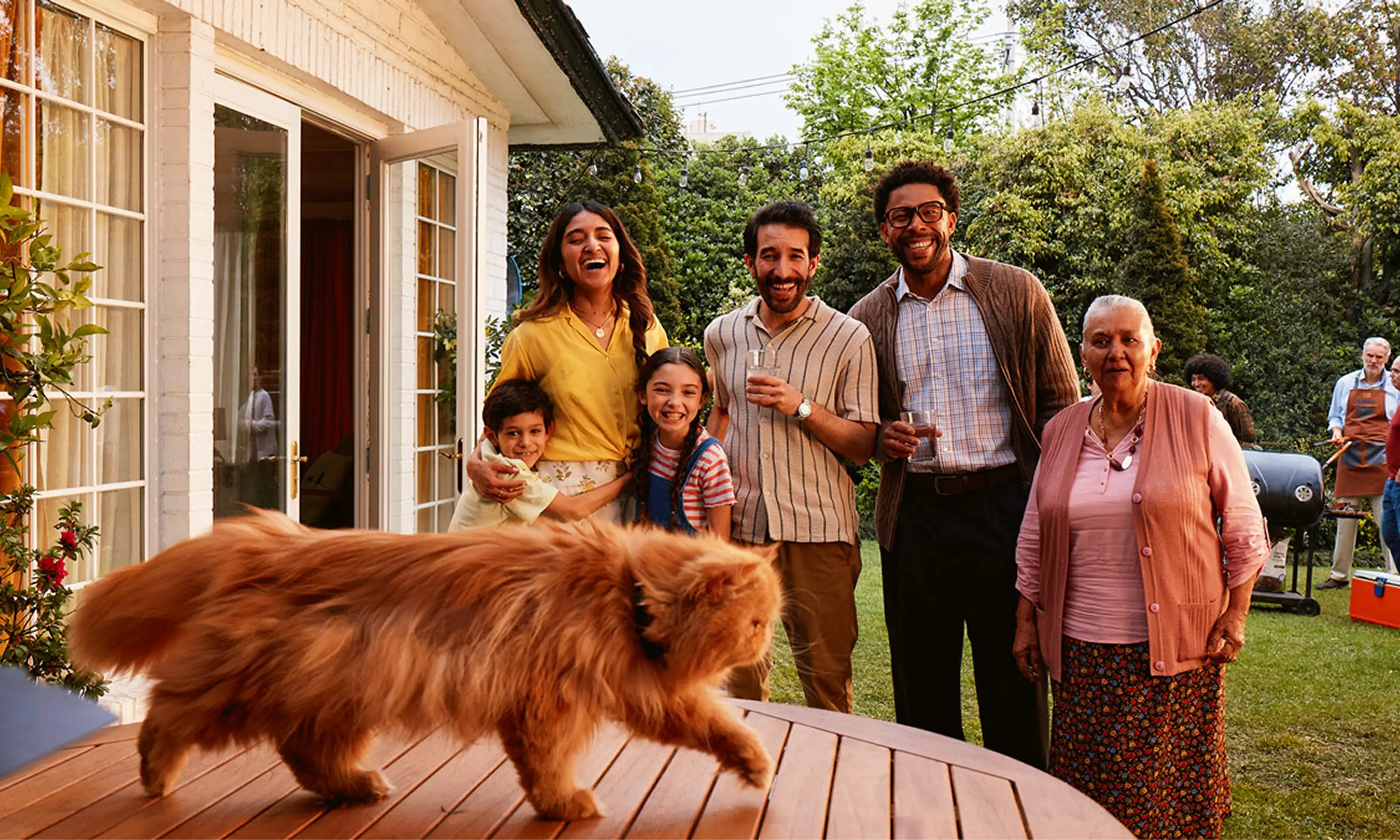 A family outside at a barbecue laughing as their cat walks across the table
