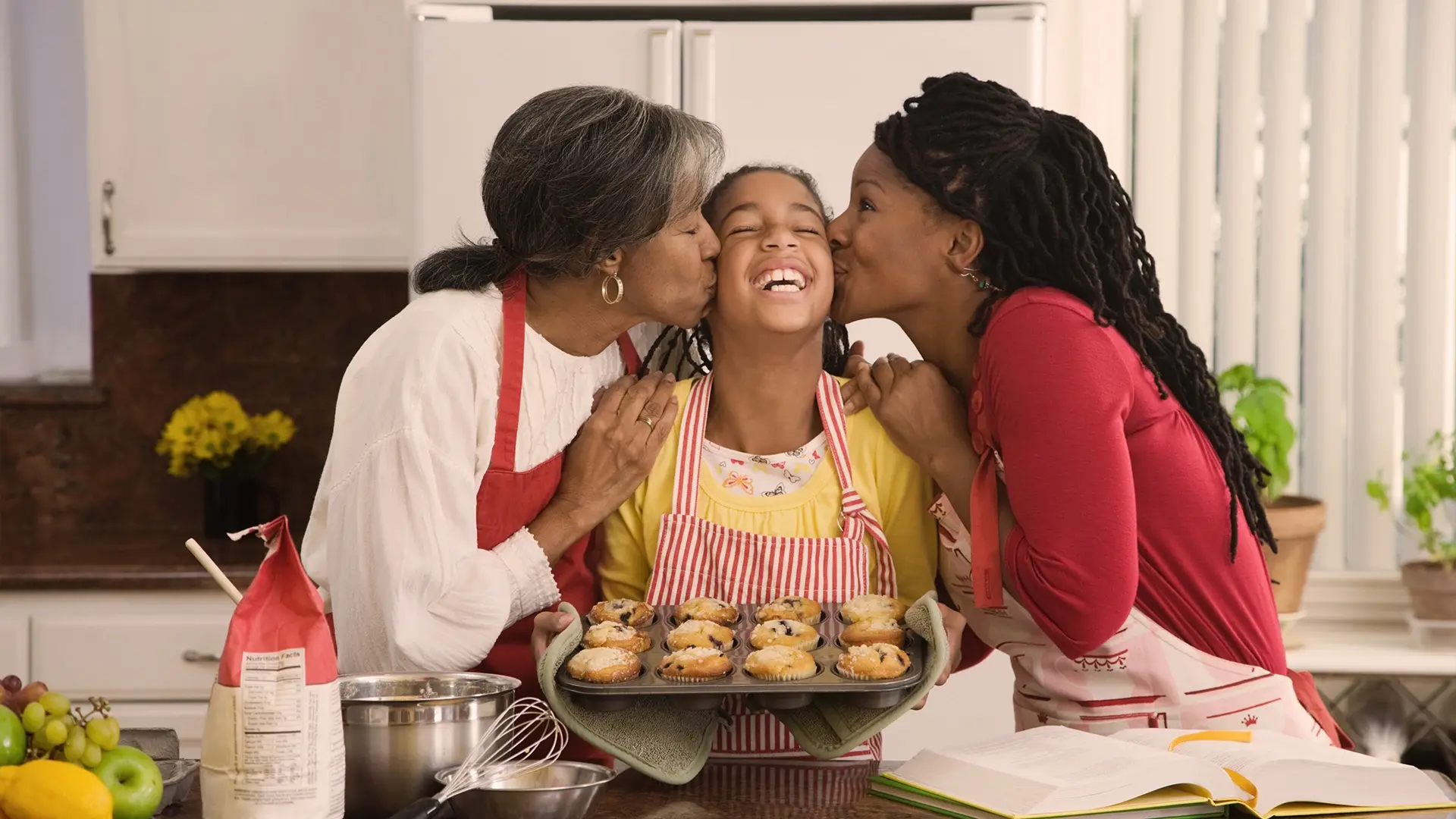 Smiling girl in a yellow shirt and striped apron holds a tray of muffins while two women kiss her cheeks in a kitchen setting.