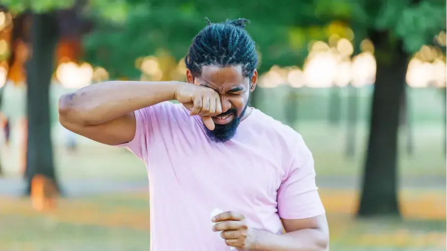 A man is outside in a short sleeved shirt, rubbing his eye with one hand and holding a tissue in the other hand.
