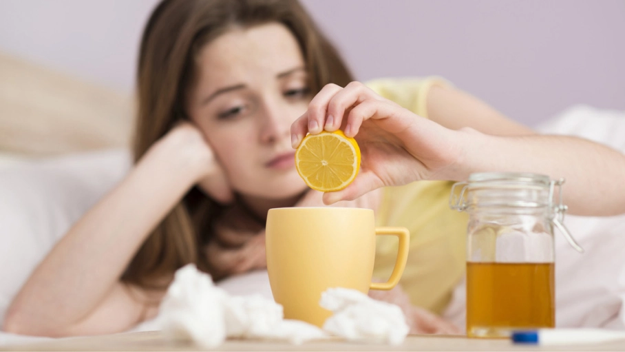 Woman, laying down, squeezing lemon into her mug next to a pile of tissues, honey and a thermometer