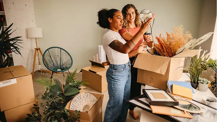 Two women unpack boxes in a room filled with plants, moving boxes, and home decor items.