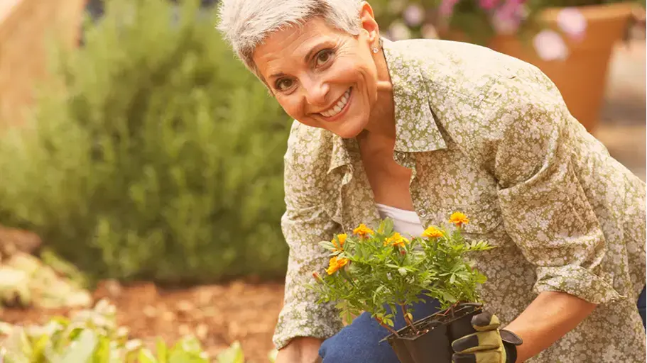 A woman smiles while holding a small potted plant in her hand as she works in her garden.