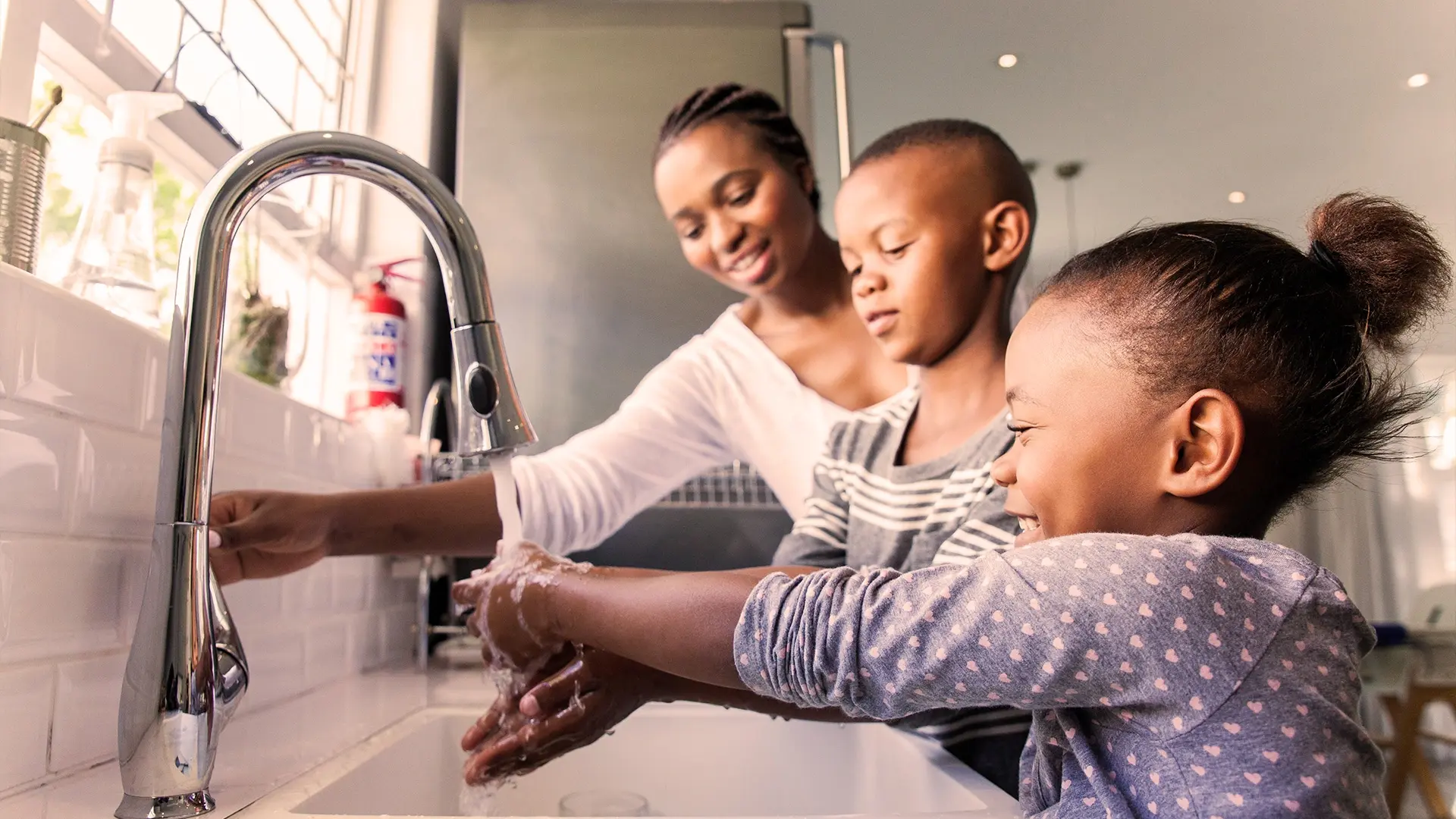Mother helps her two children wash their hands at a white kitchen sink.