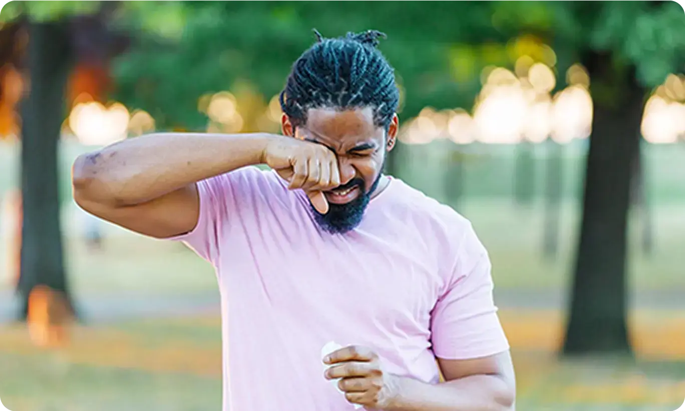 A man outdoors rubbing his eyes and nose, indicating the discomfort of outdoor allergies in a park.