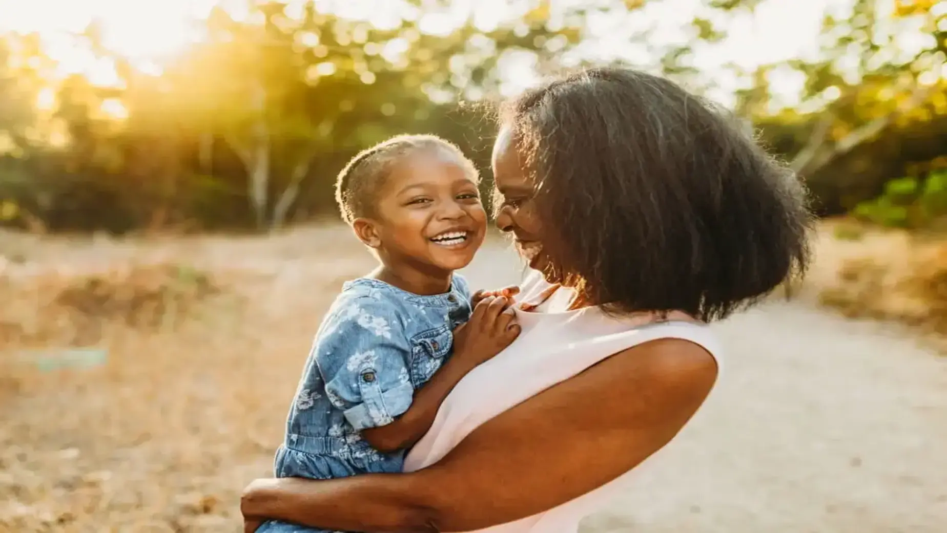 A mother holding her young child in a sunlit outdoor setting, surrounded by nature and greenery.